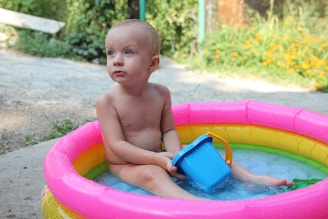 A young child is sitting in a brightly colored inflatable pool, holding a blue bucket. The pool has pink, yellow, and green rings, and it is outdoors with green foliage and flowers in the background.
