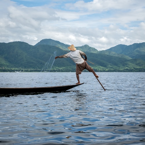 A person is practicing traditional fishing on a lake, balancing on one leg on a narrow wooden boat with a fishing net in hand. The background features a range of green hills under a partly cloudy sky.