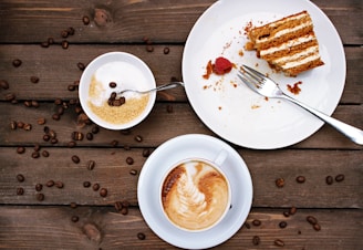 A wooden table displays a slice of layered cake with cream filling on a white plate, accompanied by a fork and a single raspberry. Next to it, a bowl contains brown sugar and a spoon with a few coffee beans on top. A cup of cappuccino with foamy milk art rests on a saucer nearby, while scattered coffee beans decorate the tabletop.