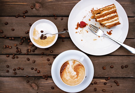 A wooden table displays a slice of layered cake with cream filling on a white plate, accompanied by a fork and a single raspberry. Next to it, a bowl contains brown sugar and a spoon with a few coffee beans on top. A cup of cappuccino with foamy milk art rests on a saucer nearby, while scattered coffee beans decorate the tabletop.