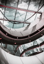 A modern architectural interior featuring a curved glass ceiling with reflections of the sky. The design includes sleek metal and glass elements with subtle red lighting accents that add contrast to the overall composition.
