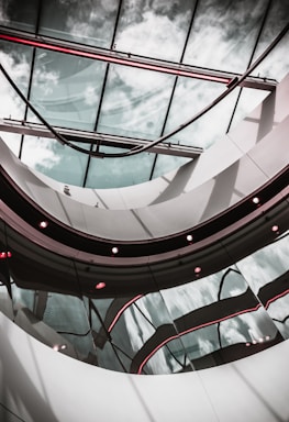 A modern architectural interior featuring a curved glass ceiling with reflections of the sky. The design includes sleek metal and glass elements with subtle red lighting accents that add contrast to the overall composition.