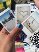 A hand with blue nail polish holds three instant photos. The first photo features a park with trees and a clear sky. The second photo displays a heart drawn in the sand. The third photo captures a person doing a cartwheel on the beach. The background includes a colorful beach towel and a polka-dotted item.