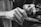 A close-up black-and-white image of a person's hands engaged in a detailed activity. One hand holds a small, pointed tool, while the other hand supports the task being performed. The focus is on the textures and movements of the hands, emphasizing precision and concentration.