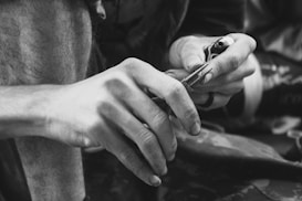 A close-up black-and-white image of a person's hands engaged in a detailed activity. One hand holds a small, pointed tool, while the other hand supports the task being performed. The focus is on the textures and movements of the hands, emphasizing precision and concentration.