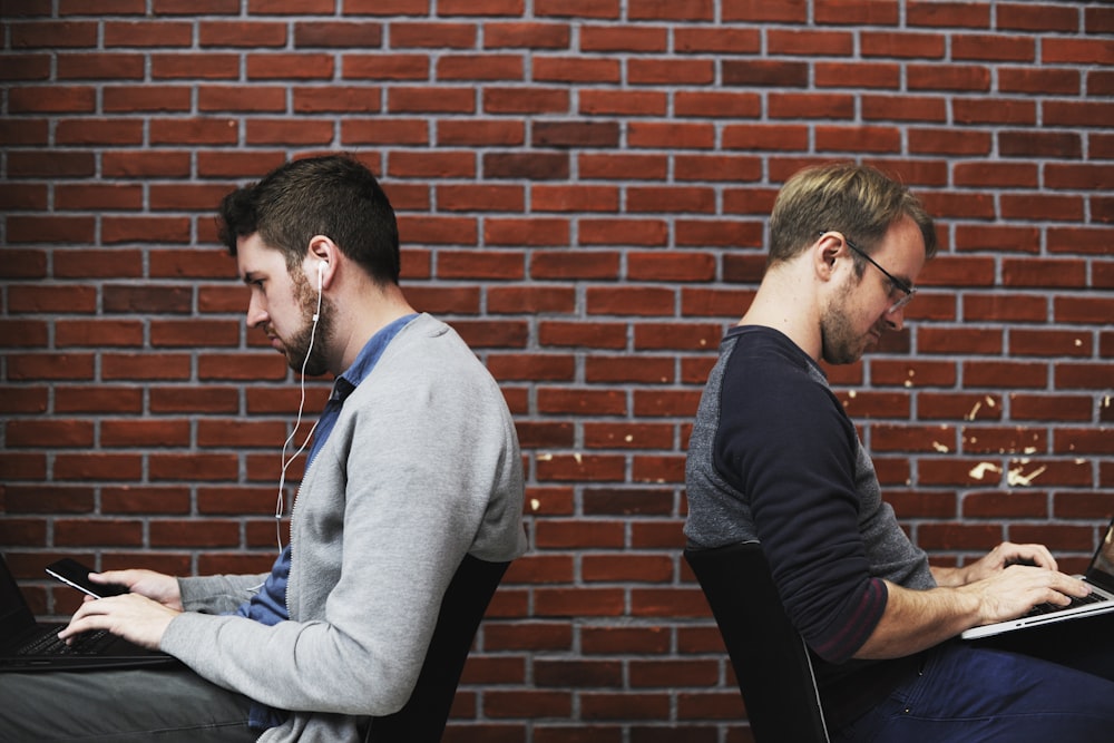 Two men working back-to-back outside with a brick wall background