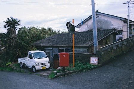 A rural scene featuring a small, rustic house with a tiled roof, surrounded by overgrown vegetation. A white mini truck is parked on the side next to a bright red mail collection box. A large convex traffic mirror is mounted on an orange pole nearby, while utility poles and wires extend against a partly cloudy sky.