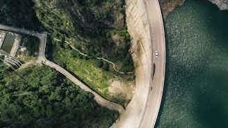 An aerial view shows a large curved dam structure next to a body of water. On the dam, a single white car is visible on the road. On the left, lush greenery and rocky cliffs are prominent, with winding pathways leading through the vegetation.