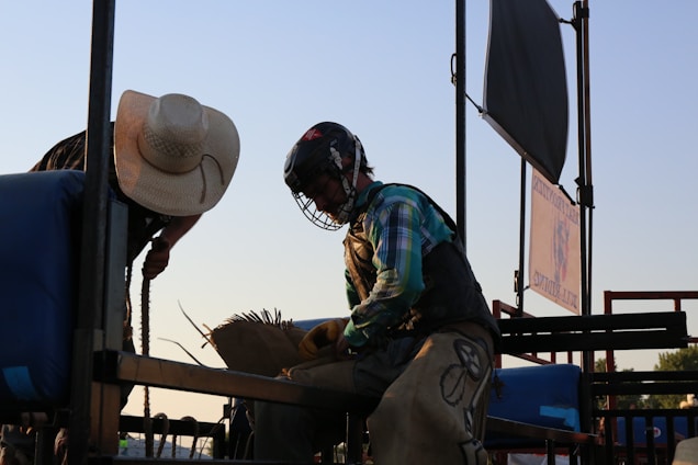 A person wearing a protective helmet and plaid shirt sits atop a rodeo bull, preparing for a ride. To the left, another individual in a straw hat leans over to assist or instruct. The setting includes metal bars and blue padding, illuminated by a sunset, creating contrasting shadows.