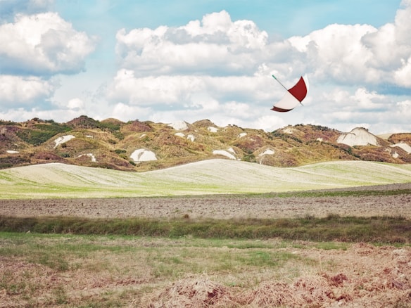 A vibrant landscape featuring rolling hills with patches of green and brown vegetation under a sky filled with fluffy white clouds. An umbrella is caught mid-air, suggesting wind, creating a surreal element against the natural backdrop.