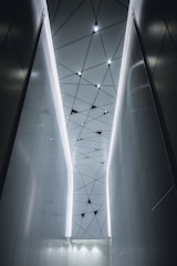 A modern interior with a geometric ceiling pattern featuring triangular shapes and integrated lights. The walls are sleek and reflective, creating a futuristic and sophisticated atmosphere.