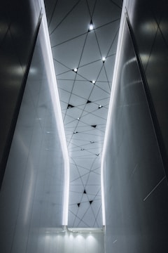A modern interior with a geometric ceiling pattern featuring triangular shapes and integrated lights. The walls are sleek and reflective, creating a futuristic and sophisticated atmosphere.