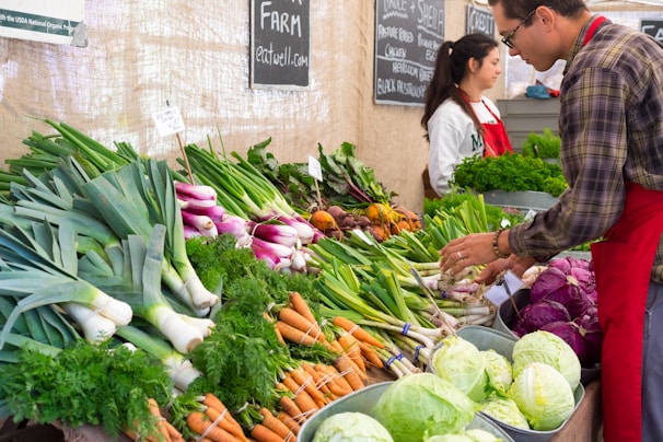 A market stall displays a variety of fresh vegetables including leeks, carrots, purple cabbages, and lettuce. Two people, a man and a woman, are tending to the produce. Handwritten signs are visible, showing the prices and names of the vegetables. The setting appears to be a busy, vibrant farmers market.