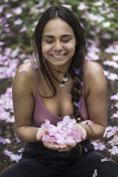 A person is sitting with eyes closed, smiling gently while holding a handful of pink flowers. The background is a soft blur of similar flowers, creating a serene and tranquil atmosphere.