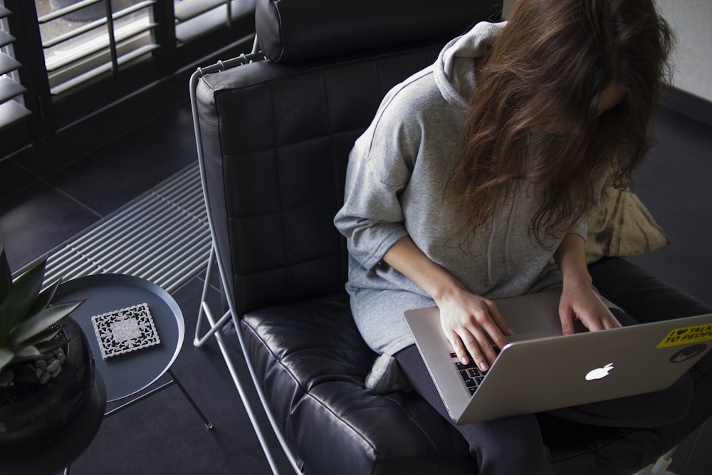 Frau in blauem Chambray-Langarmoberteil sitzt auf schwarzem Ledersessel mit silbernem MacBook auf dem Schoß