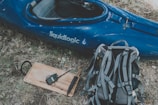A blue kayak with the word 'liquidlogic' on its side rests on dry grass next to a gray and black backpack. A wooden board with a walkie-talkie and a black object is also visible nearby.
