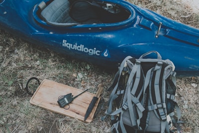 A blue kayak with the word 'liquidlogic' on its side rests on dry grass next to a gray and black backpack. A wooden board with a walkie-talkie and a black object is also visible nearby.
