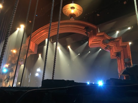The image features a stage setup with theatrical lighting. A large, ornate archway frames the background, illuminated by multiple spotlights aiming in various directions. Some stage ropes are visible in the foreground, and a hanging lamp is situated above the archway. The overall ambiance suggests a theater or concert setting, with mist or fog possibly enhancing the lighting effects.