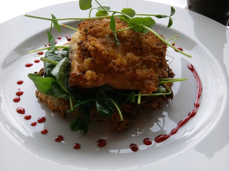 A piece of crispy, golden-brown fish is carefully plated atop a bed of greens and what appears to be slices of zucchini. The dish is garnished with pea shoots and surrounded by a decorative drizzle of red sauce on a white plate.