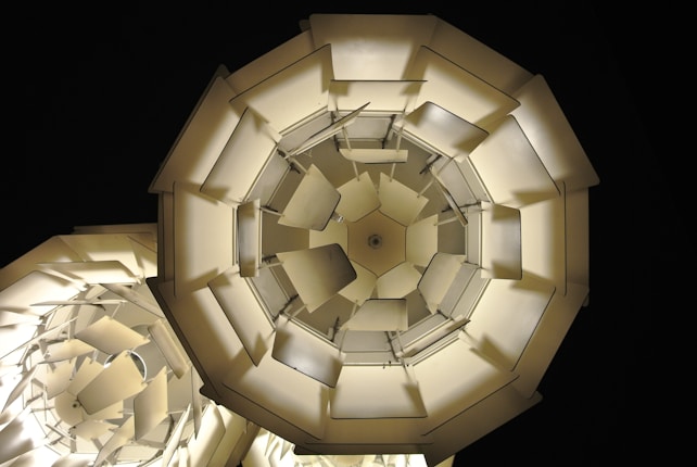 Geometric shapes with overlapping panels form intricate, layered structures that resemble abstract chandeliers or modern art installations. The light emits a warm, soft glow against a dark background, creating contrast and highlighting the complex design.