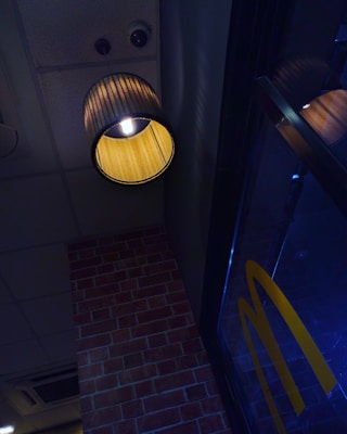 A warm-lit ceiling lamp with a cylindrical shade hangs near a brick wall. The wall features a combination of brick and glass. The glass panel reflects the interior lighting. Visible through the glass is a large yellow letter M, a familiar logo partial image.