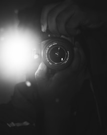 A black and white photograph captures a close-up of a person holding a camera with a lens in focus. The flash is prominently visible, causing a bright light in the image. The hands gripping the camera add an element of action and engagement.