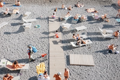 A pebbled beach with people lounging on sunbeds under a clear sky. A wooden walkway runs through the middle, providing a path among the sunbathers. Various sunbeds and towels are scattered across the beach, and individuals are either lying down, relaxing, or interacting in small groups.