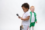 Two children are present in the image. The child in the foreground is holding a microphone and wearing a white outfit with red details. The child in the background is smiling and dressed in a green jacket and shorts with white leggings and a white shirt featuring colorful letters.