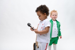 Two children are present in the image. The child in the foreground is holding a microphone and wearing a white outfit with red details. The child in the background is smiling and dressed in a green jacket and shorts with white leggings and a white shirt featuring colorful letters.