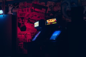 A dimly lit room features two vintage arcade game machines, one displaying 'Frogger.' The background is covered with retro-style posters, emitting a nostalgic, 80s vibe. The red and blue lighting creates an atmospheric glow.