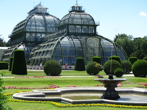 A large, ornate glass greenhouse structure stands majestically with its curved, iron frame and multiple domes. Surrounding the greenhouse are meticulously maintained gardens featuring vibrant green hedges and manicured lawns, interspersed with colorful flower beds. In the foreground, a circular fountain with water gently spraying creates a focal point in the lush setting.