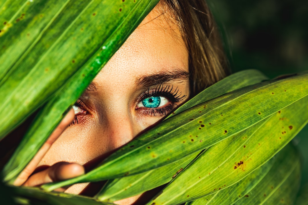 Woman with teal eye, peeking through green plant leaves.