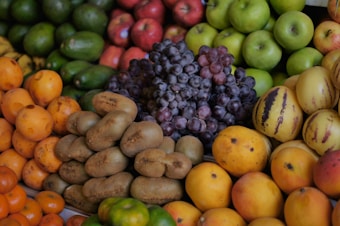 A vibrant arrangement of assorted fruits including kiwi, grapes, apples, melons, avocados, and other citrus fruits. The fruits are stacked and displayed in an inviting and colorful manner.