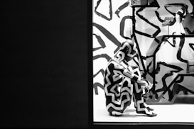 A store display features a mannequin covered in a black and white zigzag pattern. The background and surroundings also have similar abstract patterns, creating a cohesive visual theme. The mannequin is seated on a white platform with geometric shelves visible in the background.