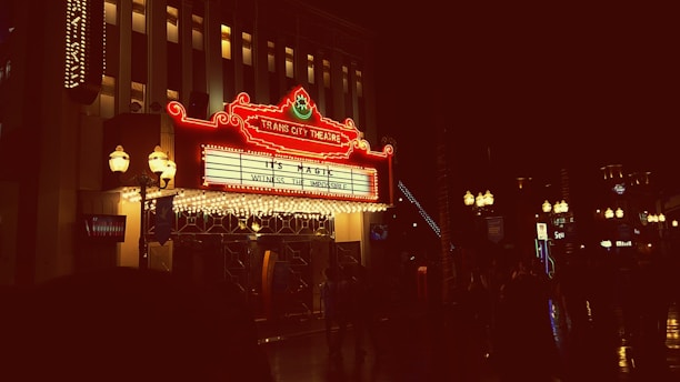 A brightly lit theater marquee displays the text 'It's Magic' and 'Witness the Impossible' at the Trans City Theatre. The neon lights illuminate the surrounding area with a warm glow, and vintage-style lamps are visible beside the entrance. Several people are seen as silhouettes walking in the dimly lit street.