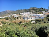 White buildings with red-tiled roofs are nestled on a hillside surrounded by lush green vegetation. The landscape extends into the mountains under a clear blue sky.
