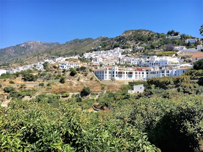 White buildings with red-tiled roofs are nestled on a hillside surrounded by lush green vegetation. The landscape extends into the mountains under a clear blue sky.