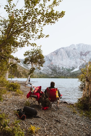 A person is sitting on a red camping chair by the side of a lake, surrounded by trees. A colorful blanket is draped over the chair, and a bag and backpack are visible on the ground. Another person stands closer to the water, fishing. Majestic mountains are visible in the background, partially covered with snow.