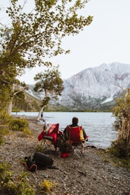 A person is sitting on a red camping chair by the side of a lake, surrounded by trees. A colorful blanket is draped over the chair, and a bag and backpack are visible on the ground. Another person stands closer to the water, fishing. Majestic mountains are visible in the background, partially covered with snow.