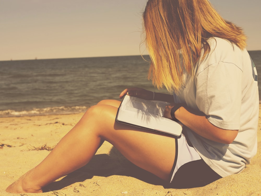 Una mujer rubia sentada en la playa mirando su libro.