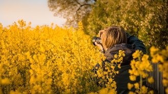 A person with short hair is standing in a field of vibrant yellow flowers, holding a camera up to their face, capturing a photograph. Lush greenery forms the backdrop, creating a peaceful and natural setting. The image is taken from behind the person, emphasizing the act of photography surrounded by nature.