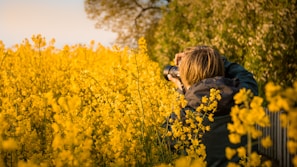 A person with short hair is standing in a field of vibrant yellow flowers, holding a camera up to their face, capturing a photograph. Lush greenery forms the backdrop, creating a peaceful and natural setting. The image is taken from behind the person, emphasizing the act of photography surrounded by nature.