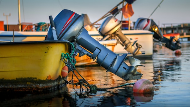 A close-up view of several motorboats docked on a calm body of water. The focus is on the metallic outboard motors extending into the water, reflecting the soft golden light of either sunrise or sunset. The boats are tied to buoys, with visible ropes and some algae growth at the waterline. The background features more boats and a hint of a pier.