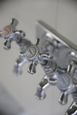 Three metallic cross-handled faucets are attached to a wall plate, each labeled with decorative text. The central faucet's label reads 'Lille Herbeau France'. The design is vintage with a polished chrome finish.