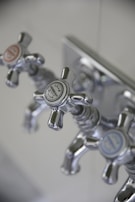 Three metallic cross-handled faucets are attached to a wall plate, each labeled with decorative text. The central faucet's label reads 'Lille Herbeau France'. The design is vintage with a polished chrome finish.