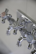 Three metallic cross-handled faucets are attached to a wall plate, each labeled with decorative text. The central faucet's label reads 'Lille Herbeau France'. The design is vintage with a polished chrome finish.