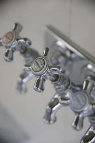 Three metallic cross-handled faucets are attached to a wall plate, each labeled with decorative text. The central faucet's label reads 'Lille Herbeau France'. The design is vintage with a polished chrome finish.