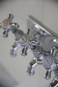 Three metallic cross-handled faucets are attached to a wall plate, each labeled with decorative text. The central faucet's label reads 'Lille Herbeau France'. The design is vintage with a polished chrome finish.