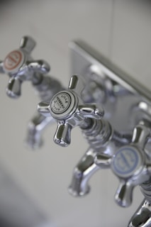 Three metallic cross-handled faucets are attached to a wall plate, each labeled with decorative text. The central faucet's label reads 'Lille Herbeau France'. The design is vintage with a polished chrome finish.