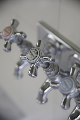 Three metallic cross-handled faucets are attached to a wall plate, each labeled with decorative text. The central faucet's label reads 'Lille Herbeau France'. The design is vintage with a polished chrome finish.